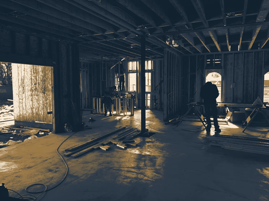 A framer in a hard hat and leather tool bags walks through a framed opening inside a Southern California house at framing stage, the bare concrete slab poured on grade underfoot and hard afternoon sun throwing the stud shadows across it in bars.